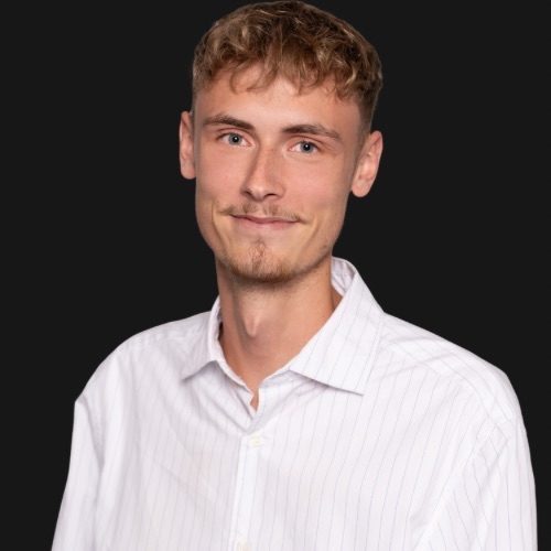 Junger Mann mit kurzem Haar, lächelnd, trägt ein helles Hemd vor schwarzem Hintergrund.