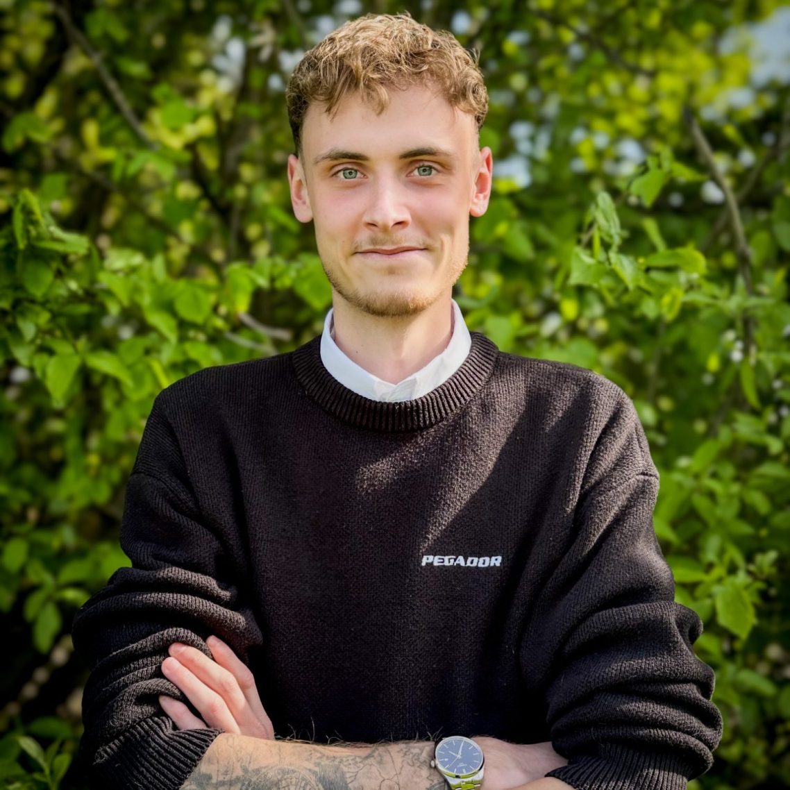 Junger Mann in schwarzem Pullover steht vor grüner Kulisse mit verschränkten Armen.