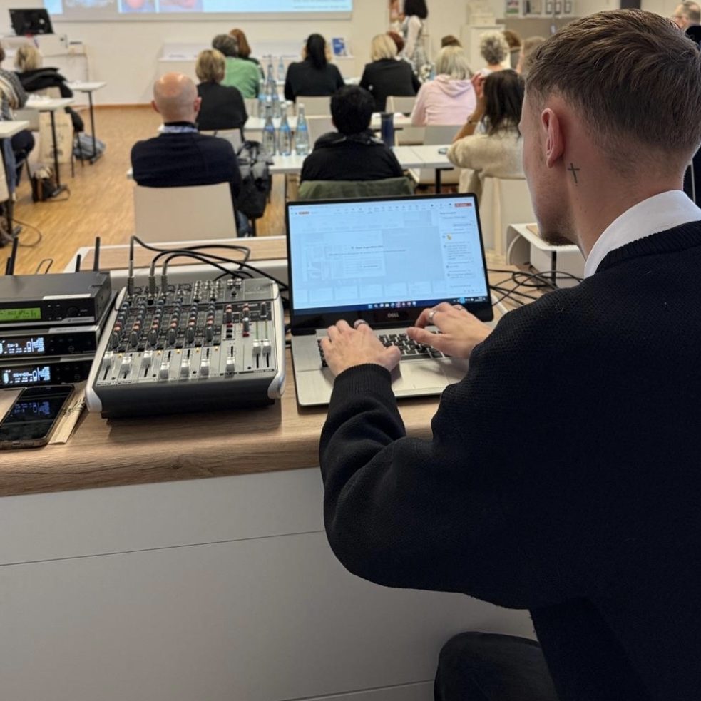 Person bedient ein Mischpult und Laptop während einer Präsentation im Hintergrund.