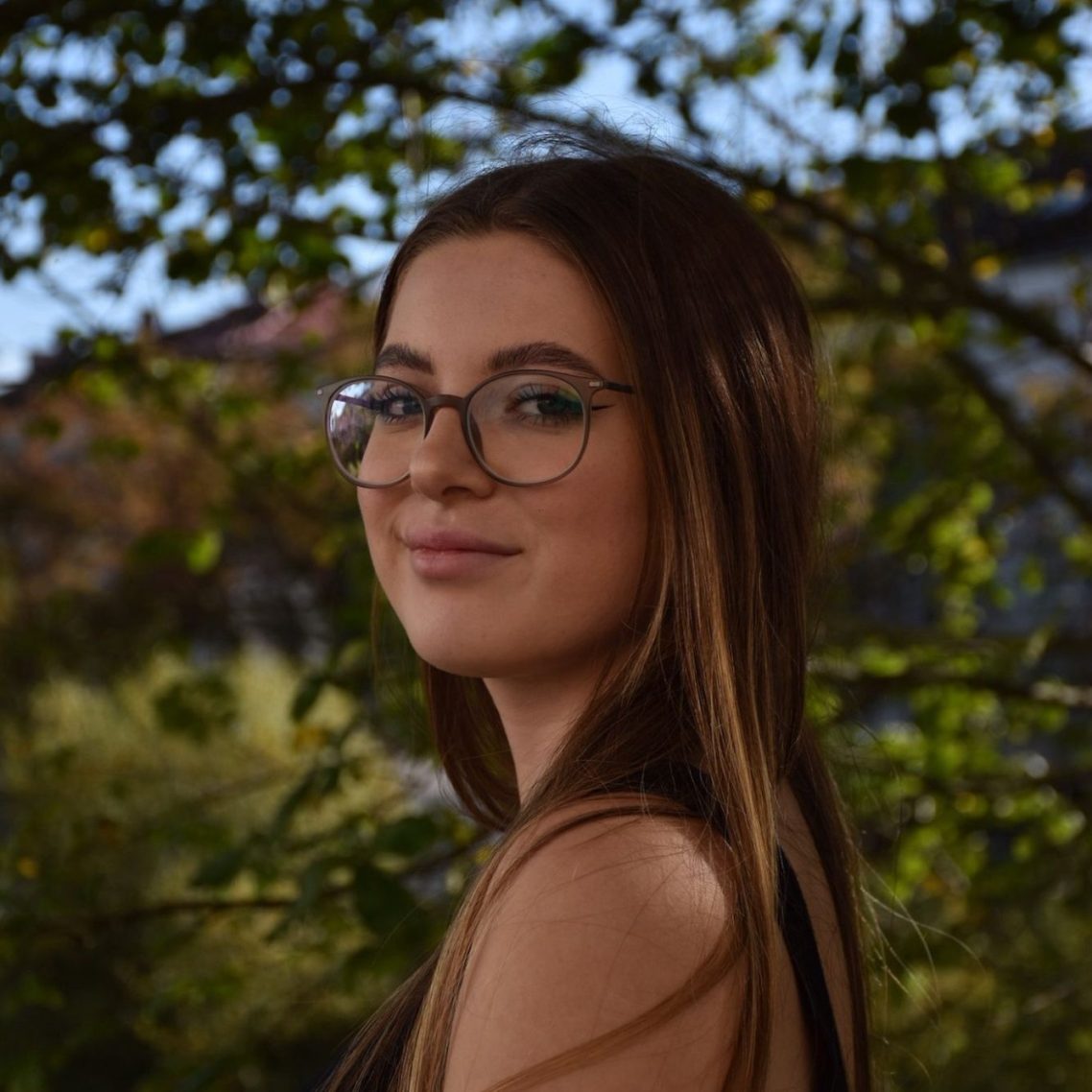 Junge Frau mit langen Haaren und Brille, steht im Freien vor Bäumen.