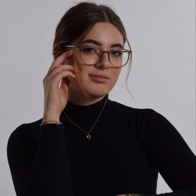 Junge Frau in schwarzem Rollkragenpullover, trägt Brille und schaut selbstbewusst.