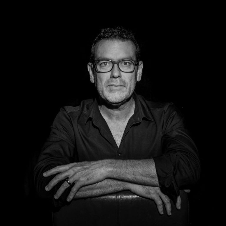 Ein Mann mit Brille in Schwarz-Weiß, in einer nachdenklichen Pose vor schwarzem Hintergrund.